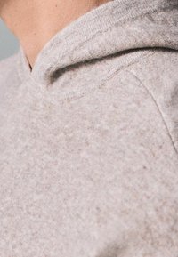 Zbliżenie osoby w jasnobeżowym, miękkim swetrze dzianym z złożonym kołnierzem.