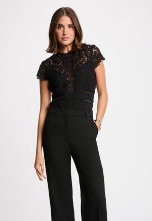 Femme portant un haut en dentelle noire à manches courtes et un pantalon noir taille haute, debout avec une main dans la poche contre un fond uni.