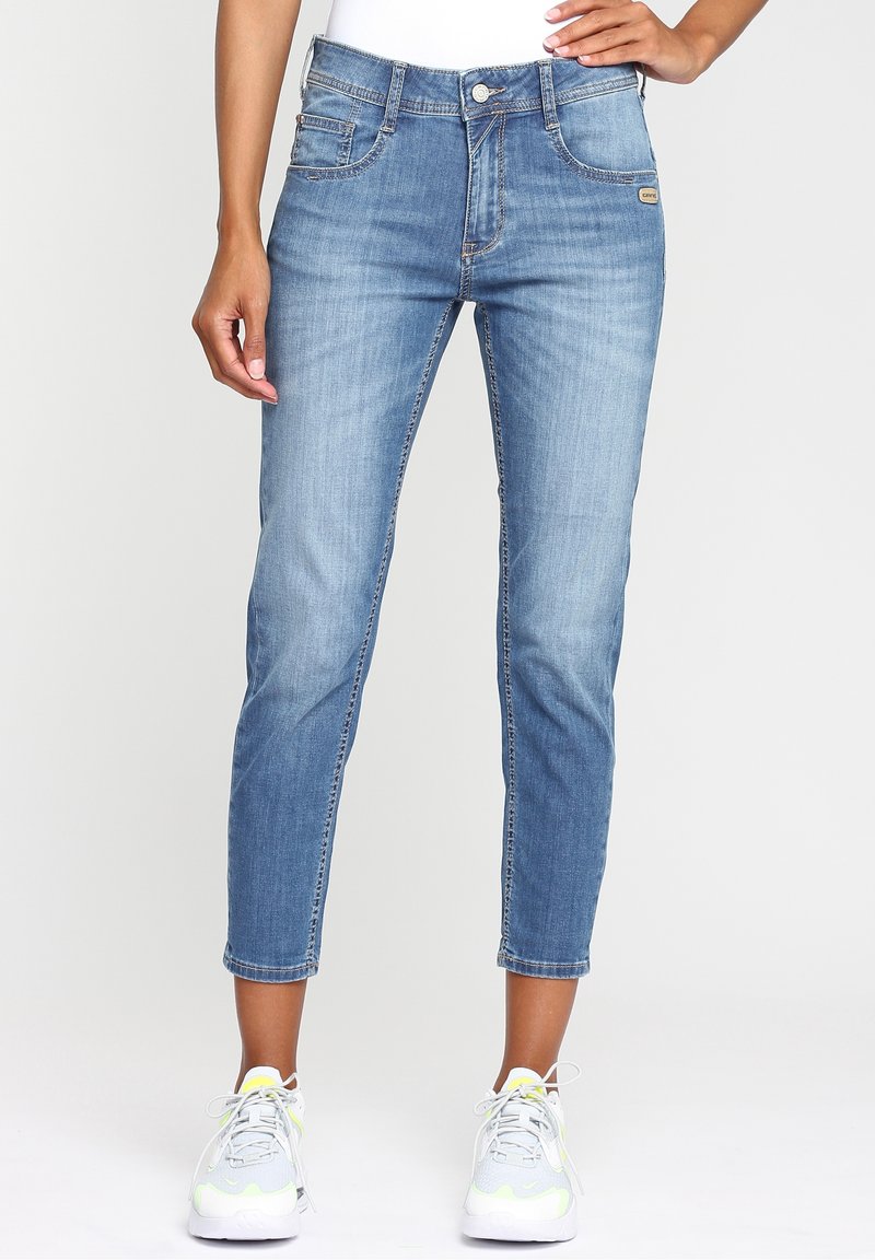 Kvinna som bär ljusblå avkortade skinny jeans och vita sneakers, stående med ena handen vilande på höften.