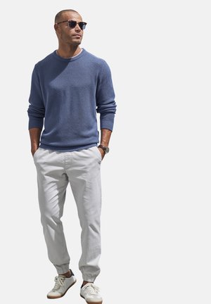 Blauer Strickpullover mit rundem Halsausschnitt, hellgraue elastische Hosen mit Bündchen und weiße Turnschuhe. Sleek Sonnenbrillen und eine Uhr ergänzen das Outfit.