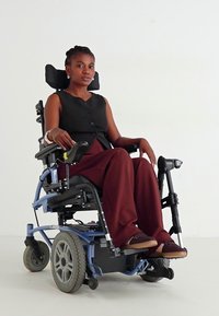 Ηλεκτρικό αμαξίδιο σε μπλε χρώμα με μεγάλους τροχούς; ο χρήστης φοράει μια μαύρη γιλέκα και μπορντό παντελόνι; καθιστή στάση, με έντονη έκφραση.