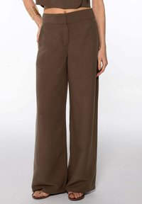 Femme portant un pantalon marron taille haute et large associé à un top court, des bracelets en or et des sandales ouvertes marron.