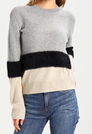 Vrouw draagt een geblokte trui in grijs, zwart en beige met een blauwe spijkerbroek, staand tegen een neutrale achtergrond.