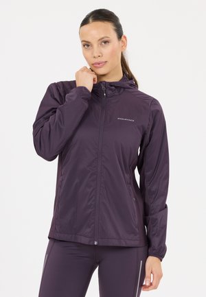 Kvinde iført en mørkelilla sportstrøje med lynlås og "Endurance" logo, kombineret med matchende leggings, stående mod en ensfarvet hvid baggrund.