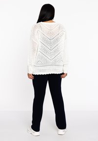 Weißer Strickpullover mit Chevron-Muster, locker sitzend, mit langen Ärmeln und gewelltem Saum. Kombiniert mit dunklen Hosen und Sneakers.
