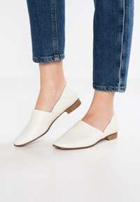 Chaussures blanches en cuir sans lacets avec un bout pointu et un petit talon en bois, portées avec un jean en denim retroussé. Texture lisse et design simple.