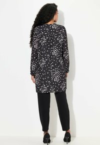 Czarna tunika z długim rękawem w białe plamki, o asymetrycznym dole i plisowanym tyłem, zestawiona z dopasowanymi czarnymi spodniami i eleganckimi butami na obcasie.