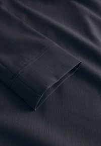 Sötétszürke hosszú ujjú ruhadarab ujja, sima textúrával és finom bordázott mintázattal. Az ujja mandzsettája tiszta kivitelű.