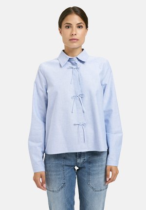 Frau in einer hellblauen Bluse mit langen Ärmeln und drei vorderen Bindebändern, kombiniert mit ausgebleichten blauen Jeans, steht vor einem weißen Hintergrund.