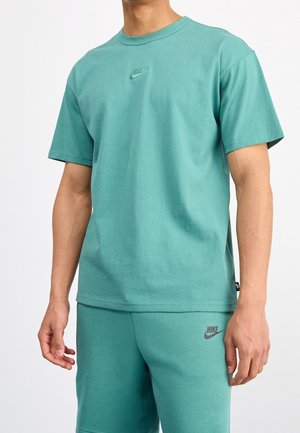 Hombre vistiendo una camiseta Nike color teal y pantalones cortos a juego con logotipos en el pecho y el muslo, de pie contra un fondo blanco liso.