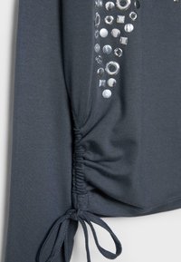 Ciemnoszara bluzka z długim rękawem, marszczonym szwem bocznym oraz srebrnymi metalicznymi okrągłymi i kwadratowymi ćwiekami w okolicy ramienia.