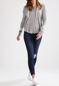 Sudadera gris con cordones blancos, de manga larga y rayas blancas en los puños, combinada con jeans skinny azul marino y zapatillas blancas.