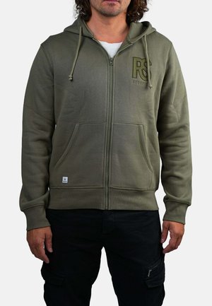 Homme portant un hoodie zippé vert olive avec des poches avant et un pantalon cargo noir, se tenant contre un fond blanc.