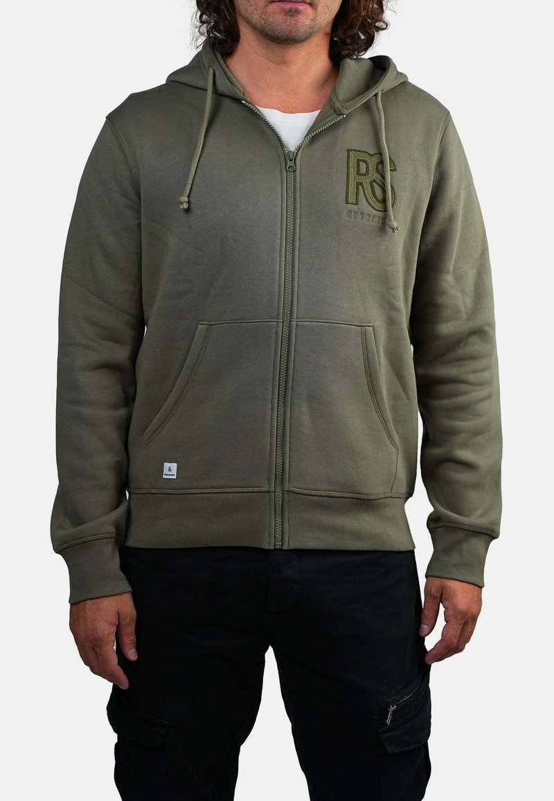 Homme portant un hoodie zippé vert olive avec des poches avant et un pantalon cargo noir, se tenant contre un fond blanc.