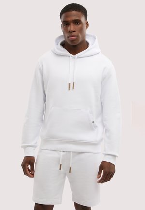 Młody mężczyzna w białej bluzie z kapturem i kieszenią z przodu oraz białych szortach ze sznurkiem, stojący na jasnym tle.