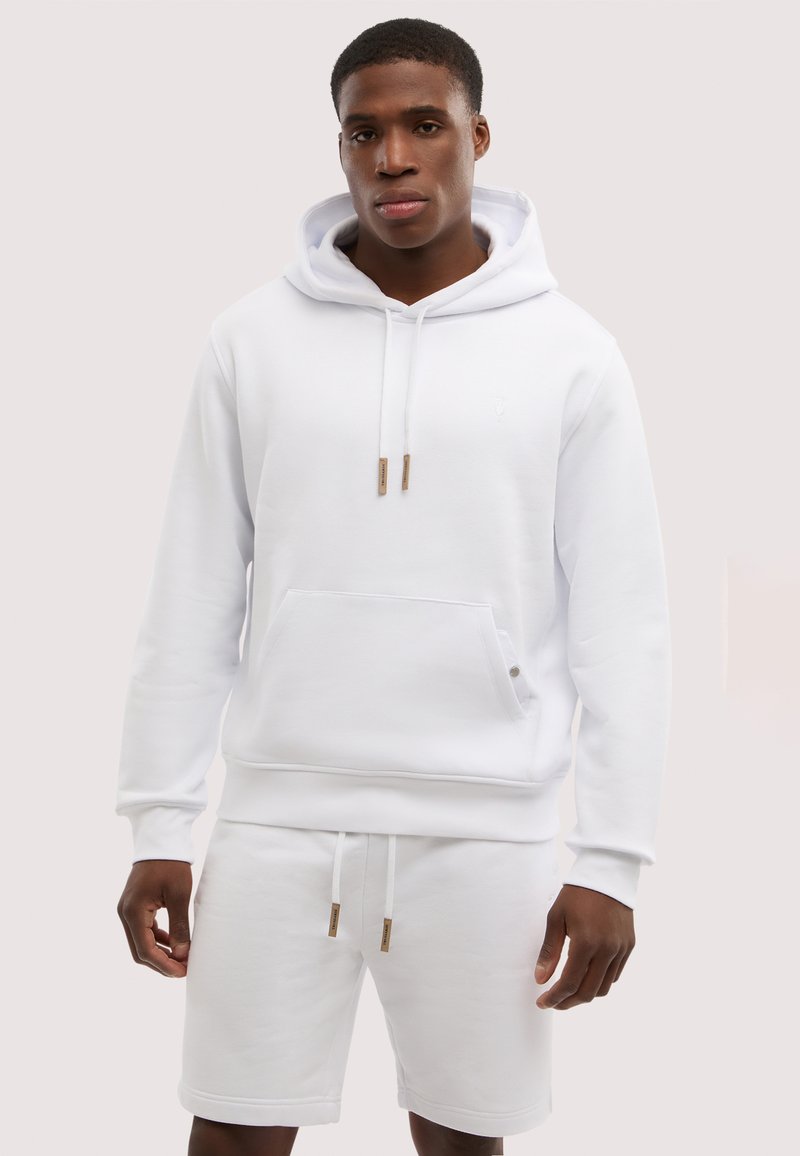 Joven con sudadera blanca con capucha y bolsillo frontal, y pantalones cortos blancos con cordón, de pie frente a un fondo claro.