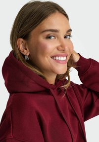 Hoodie bordeaux con un grande cappuccio, tasca frontale e coulisse. Texture del tessuto liscia, modello sorridente con orecchini e mano nei capelli.