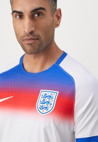 Hvid atletisk skjorte med blå og rød gradient, der har et tekstureret stof, en blå krave og et broderet England-våbenskjold.