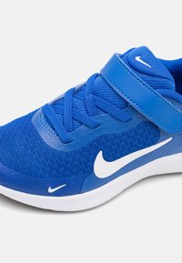 Baskets Nike bleues en tissu mesh, avec des lacets et une sangle Velcro sur une semelle blanche, sur fond clair uni.