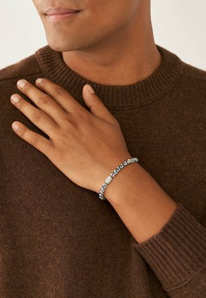 Bracelet en argent et doré avec des maillons de chaîne entrelacés et un accent rectangulaire texturé. Porté sur une main contre un pull marron.