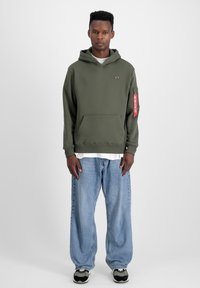 Felpa verde oliva con cappuccio e tasca a marsupio, strato interno bianco e tasca con zip sulla manica. Abbinata a jeans blu larghi e sneakers.