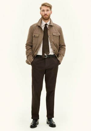 Veste en tissu brun avec des poches sur la poitrine, portée sur une chemise blanche et une cravate brune. Un pantalon noir et des chaussures complètent la tenue.