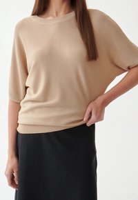 Maglione beige lavorato a maglia con maniche corte, collo tondo e orlo a coste, abbinato a una gonna nera. Tessuto morbido, vestibilità comoda.