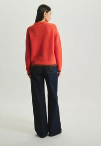 Pull en orange à côtes avec un col rond et un ourlet marron, associé à un jean bleu foncé à jambe évasée. La texture semble douce et fluffy.