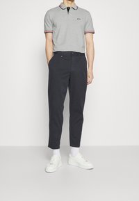 Grå polo skjorta med svart krage och röda detaljer, kombinerad med mörka byxor och vita sneakers. Modellen står mot en enhetlig vit bakgrund.