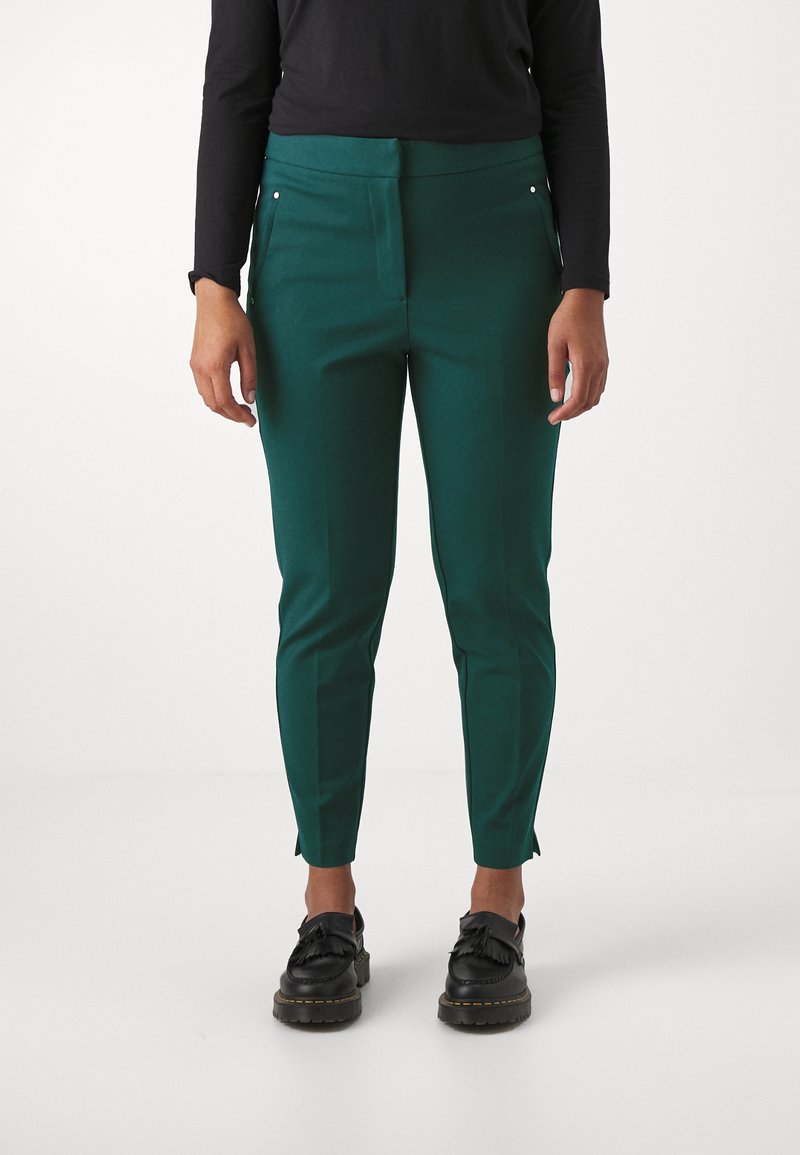 Green tailored pants with a smooth texture, featuring side pockets, a sleek silhouette, and subtle split hems at the ankle. Paired with black shoes.