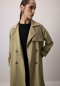 Bež plašč s širokimi reverji, dvojno zapenjanje in gumbnimi detajli. Mehka tkanina, dolgi rokavi z zaponkami.