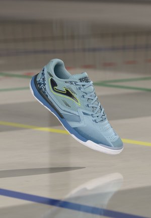 Scarpa sportiva da interno azzurro chiaro con logo nero e giallo neon, tomaia in mesh e suola bianca sospesa sopra un pavimento da palestra.