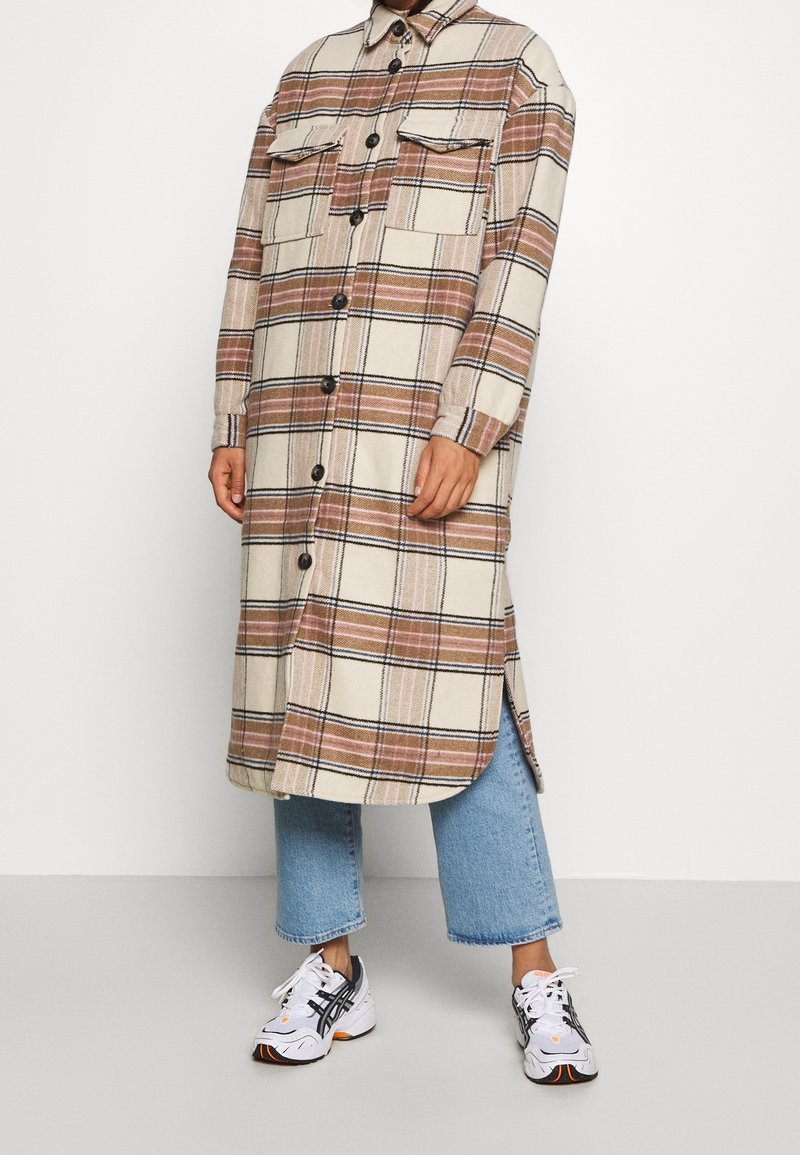 Personne portant un manteau long beige et marron à carreaux avec boutons et poches poitrine, jean bleu court et baskets blanches.