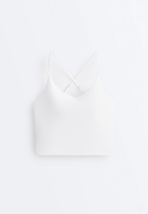 Mladá žena stojaca čelom v bielej krátkej tielkovej blúzke, čiernych legínach s vysokým pásom a bielych teniskách na jednoduchom pozadí.