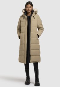 Sieviete, tērpusies garā bēžā pufīgā mētelī ar kažokādas oderi, melnās legingās un melnās kurpēs ar auklēm, stāv ar rokām kabatās.