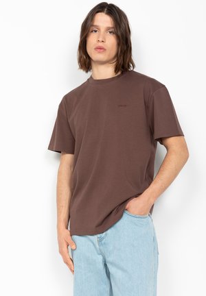 Jeune personne aux cheveux bruns mi-longs, portant un T-shirt marron uni et un jean bleu clair, debout, une main dans la poche.