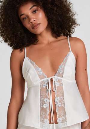 Vrouw met krullend haar die een witte satijnen en kanten camisole draagt met bloemrijke borduursels en een strikdetail aan de voorkant