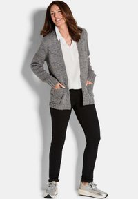 Frau, die lächelt und einen grauen Strickcardigan, eine weiße Bluse, schwarze Hosen und silberne Sneakers trägt, steht mit den Händen in den Taschen des Cardigans.