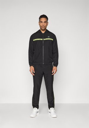 Homme portant un survêtement noir Lacoste avec une bande logo jaune fluo et des baskets blanches, sur fond clair.