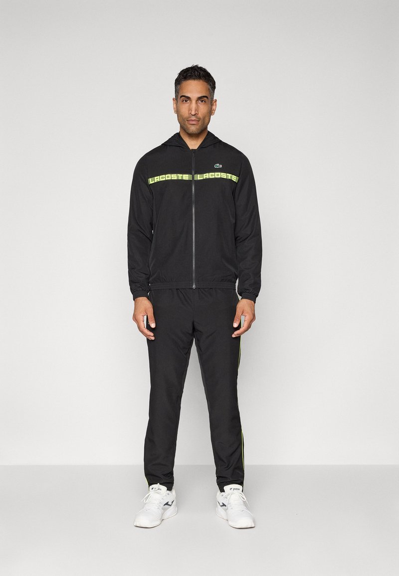 Mężczyzna w czarnej dresowej odzieży Lacoste z neonowo żółtym logo i białych sneakersach na jednolitym jasnym tle.