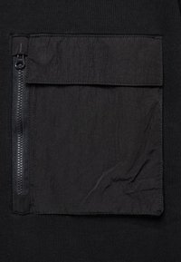 Poche en tissu noir avec rabat et fermeture éclair verticale noire sur le côté gauche, fixée à un fond textile noir.