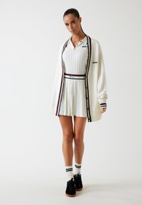 Abito bianco a coste con gonna plissettata, sovrapposto a un cardigan bianco con bordo a righe. Scarpe nere e calze al ginocchio completano il look.