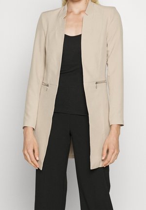 Mujer con blazer largo beige con bolsillos con cremallera sobre top negro y pantalones negros contra un fondo liso.