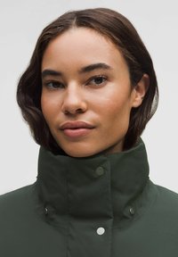 Tmavě zelená parka s vysokým límcem, se stříbrnými patentkami, texturovanou látkou a těsným střihem. Výrazné švy kolem límce.