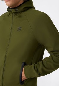 Persoon in olijfgroene zip-up hoodie met capuchon, zwarte ritsdetails en het Champion-logo op de borst en mouw, hand in de zak.