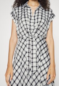 Košilové šaty s knoflíkovým zapínáním, šedým a černým kostkovaným vzorem, krátkými rukávy a páskem v pase. Hladká, lehká textilie.