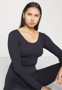 Svart ribbad långärmad topp med rundad halsringning. Tillverkad av ett elastiskt material, har den en figurnära silhuett och en slät textur.