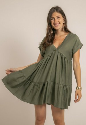 Femme souriante, portant une robe courte vert olive à col en V avec une jupe à volants, tenant le côté de la robe, accessoirisée avec des boucles d'oreilles et un bracelet dorés.