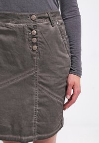 Bruine katoenen rok met knoopsluiting, voorzien van vijf decoratieve knopen, een zijsplit en zichtbare stiksel details.