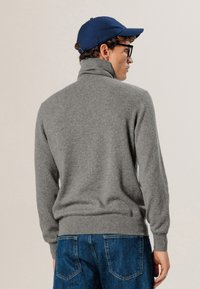 Grau gestrickter Pullover mit hohem Kragen, gerippten Bündchen und Saum. Kombiniert mit blauen Denim-Jeans, von hinten betrachtet.